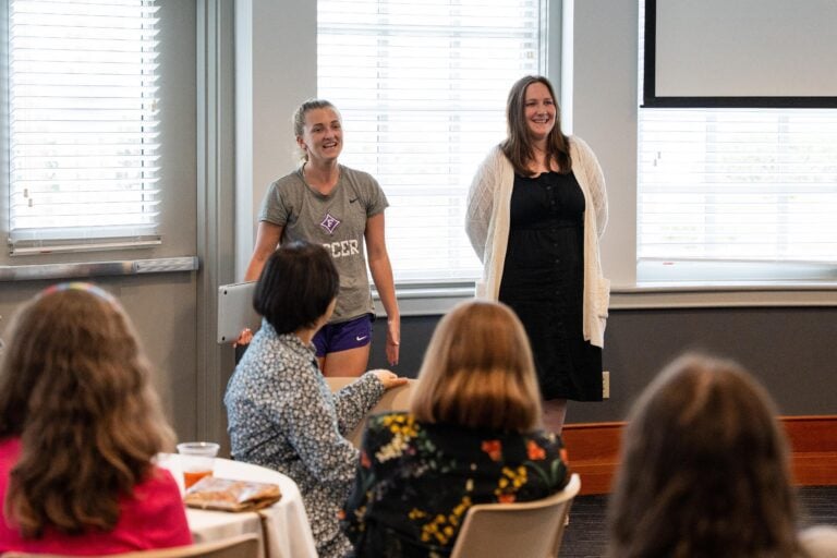 Two young women stand in front of a crowd, giving a presentation in a well-lit room.