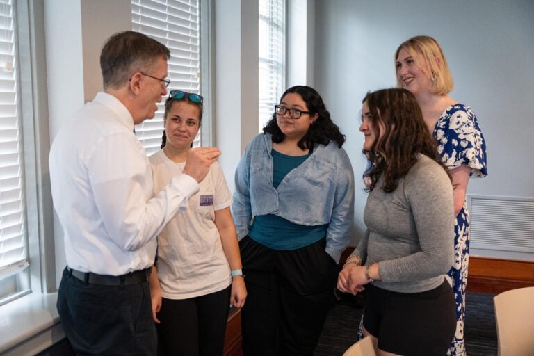 Four young women speak with an older man who is a professor following an event in a well-lit room.