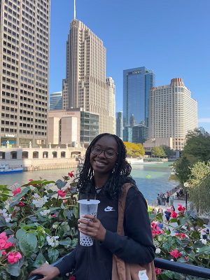 A Black woman holding a tall cup wears a dark top. She poses in front of flowers with a city skyline in the background.