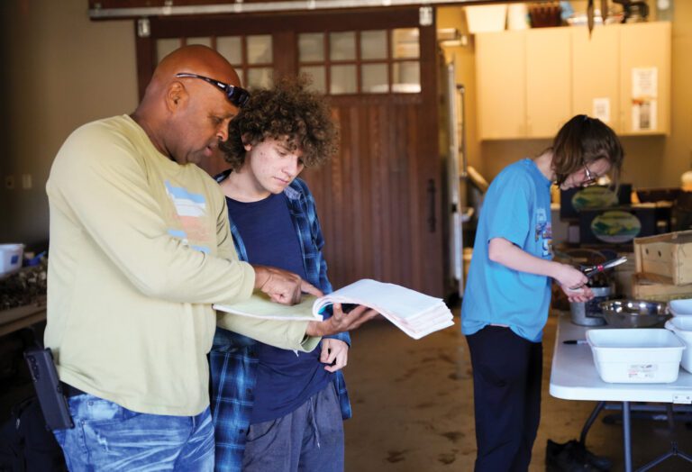 A man shows a male student something in a large notebook in front of a large wooden sliding door with small square window panes at the top.