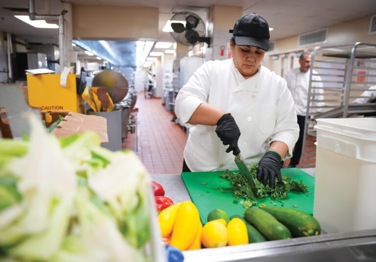A female chef chops kale and other vegetables on a green cutting board in an industrial kitchen.