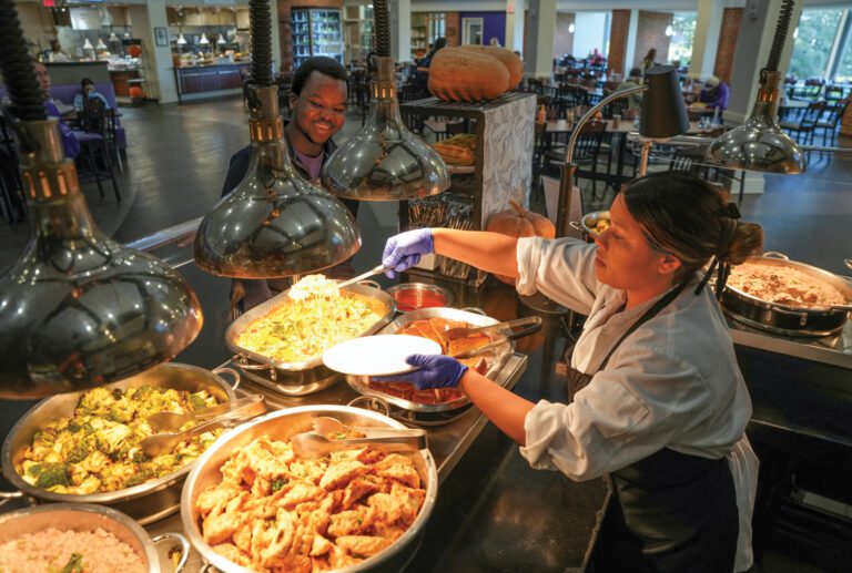 A woman serves food buffet style to students in a dining hall.