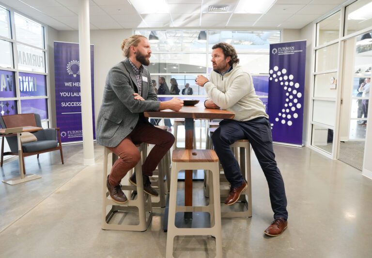 Two men have a discussion at a high top table with stools inside a bright white room with purple banners.