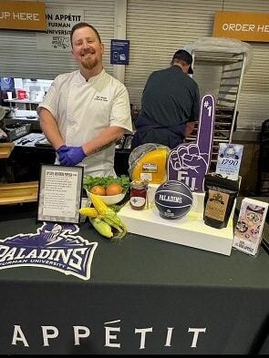A white man in a chef's coat wears food prep gloves and stands behind table of ingredients and school swag.