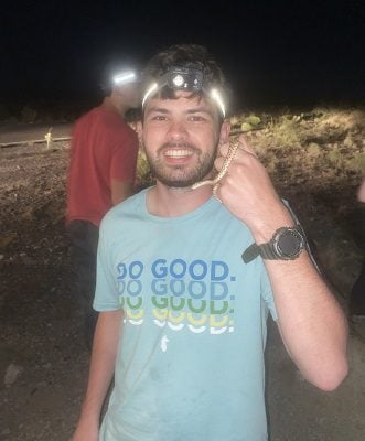 A white male with facial hair wears a headlamp and a blue T-shirt. He holds a small snake.