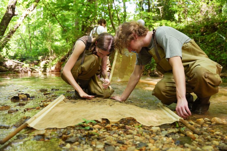 A young woman and young man squat in a muddy creek bank, looking at a net for sifting through river silt.