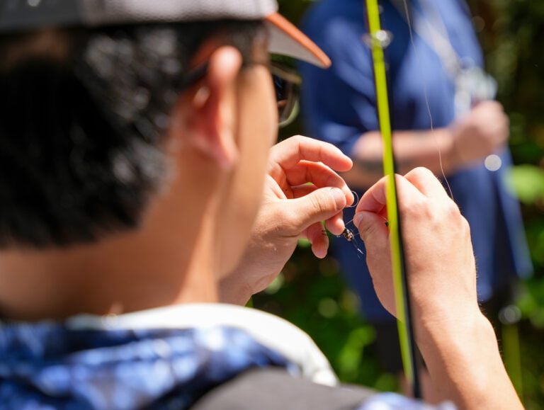 The back of a man's head as he ties a fly fishing lure onto a line.