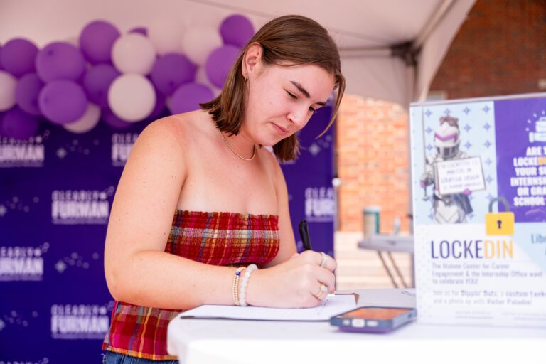 A young woman stands at a waist-high table, signing her name on a piece of paper