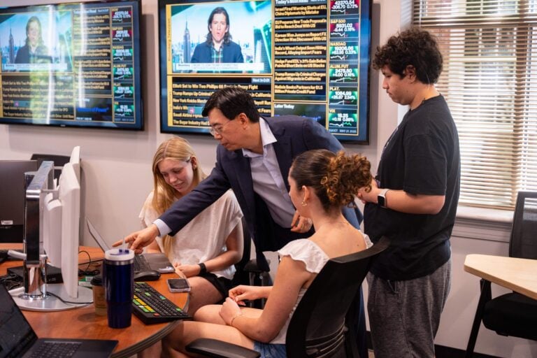 A male instructor points to a computer screen while two female students and one male student look at the screen. There are financial programs on a television in the background.