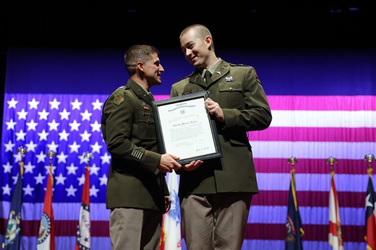 Two men dressed in green Army uniforms stand on stage in front of a large flag.