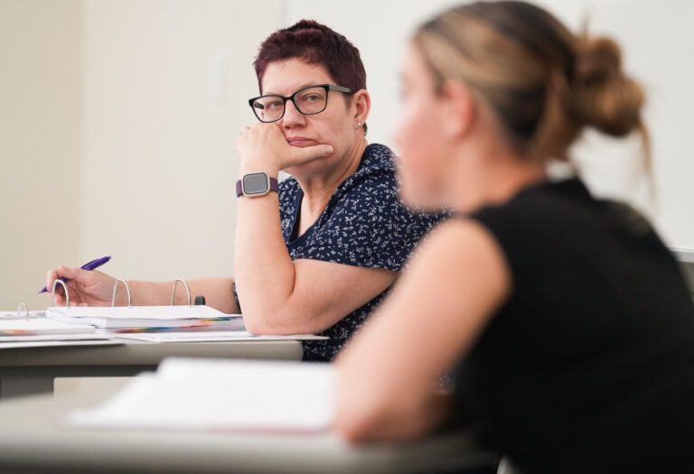 A short-haired woman sits, resting her head on her chin as she looks at a seated young woman in the foreground and takes notes on her performance in a law class.