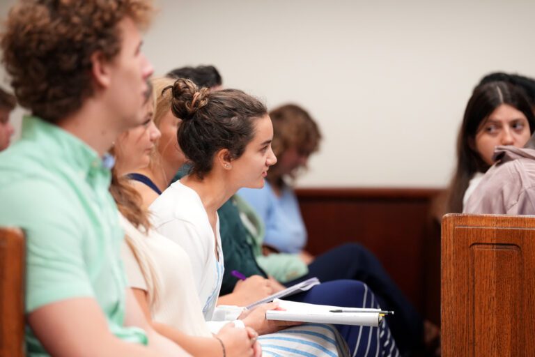 A young white woman leans forward while seated beside other people in a wooden pew in a courtroom.