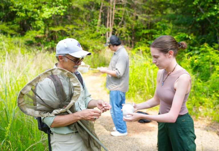 An older man and younger woman look at an insect in a jar while standing outdoors.