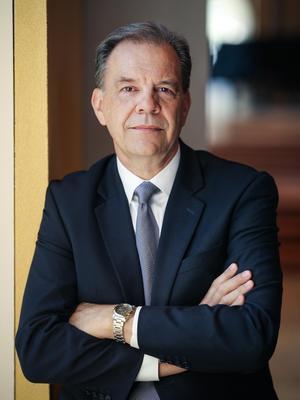 A white man in a dark suit leans against a wall with arms crossed.