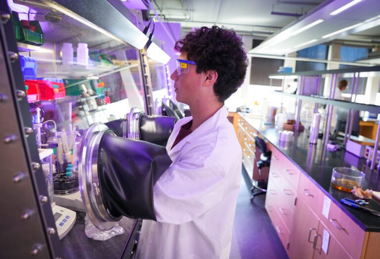 A young man in a lab coat has his arms inserted into the sleeves of a fume hood in a chemistry lab, manipulating containers of chemicals within the sealed fume hood.