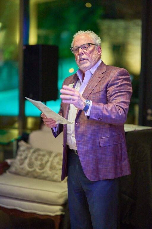 A man stands holding a microphone while wearing a purple suite and gesturing to people he is speaking to, who are not pictured