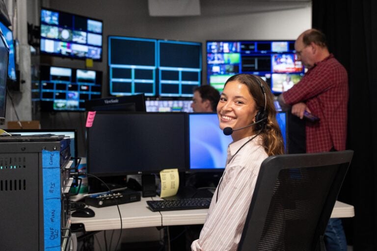 A young woman with a headset microphone on smiles seated in a television news production room.