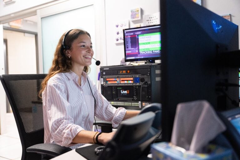 A young woman on an internship with a headset on sits at a desk in front of a computer in a television news production room.