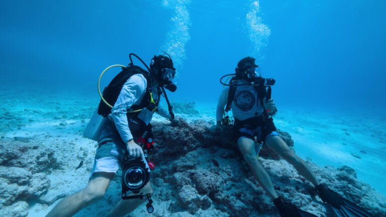 Two men in scuba gear sit on the ocean floor holding cameras and looking around