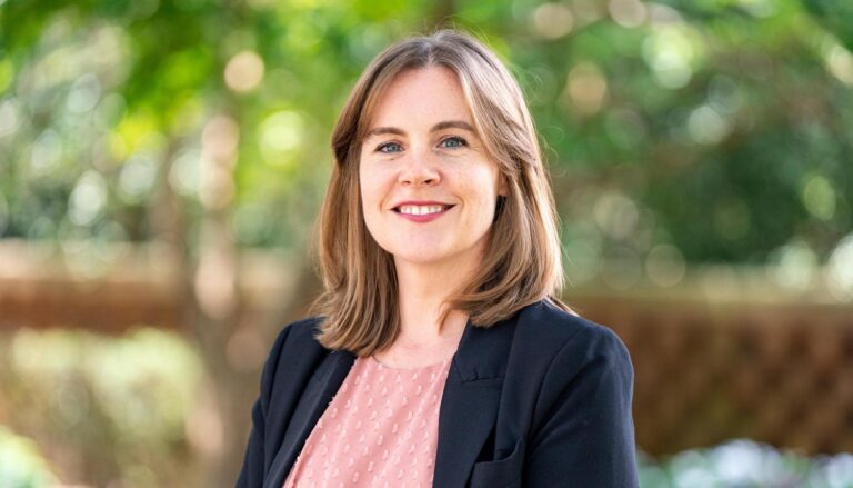 A white woman with shoulder-length hair wears a pink shirt and a dark blazer in an outdoor photo, Kathleen Casey.