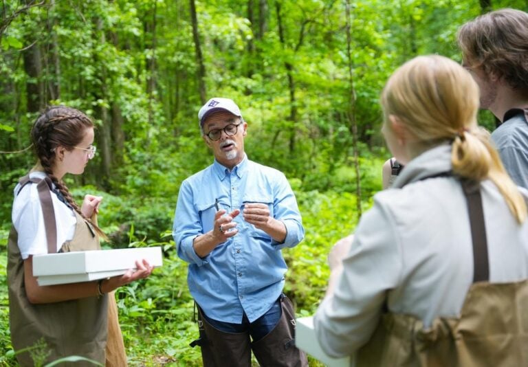 Students stand around a faculty member who's holding something in his hand. They're outside in the woods.