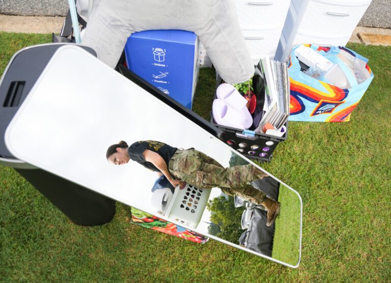 A shot of a woman's reflection in a mirror laid against a pile of belongings on a lawn. The woman is dressed in military fatigues as she helps carry luggage on Furman University's move-in day.
