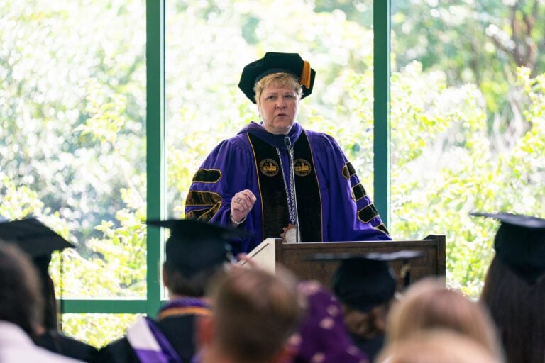 A white woman dressed in a purple robe and black ceremonial hat, the president of Furman University, stands behind a lectern addressing a crowd at a summer commencement ceremony.