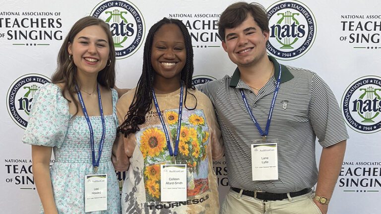 Three young people, a white woman, a Black woman and a white man, stand in front of a banner and smile at the camera.