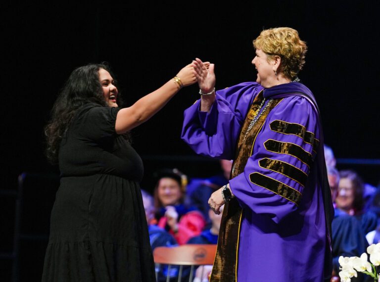 A young woman and an older woman in an academic robe share a secret handshake on stage at a Convocation ceremony for Furman University.
