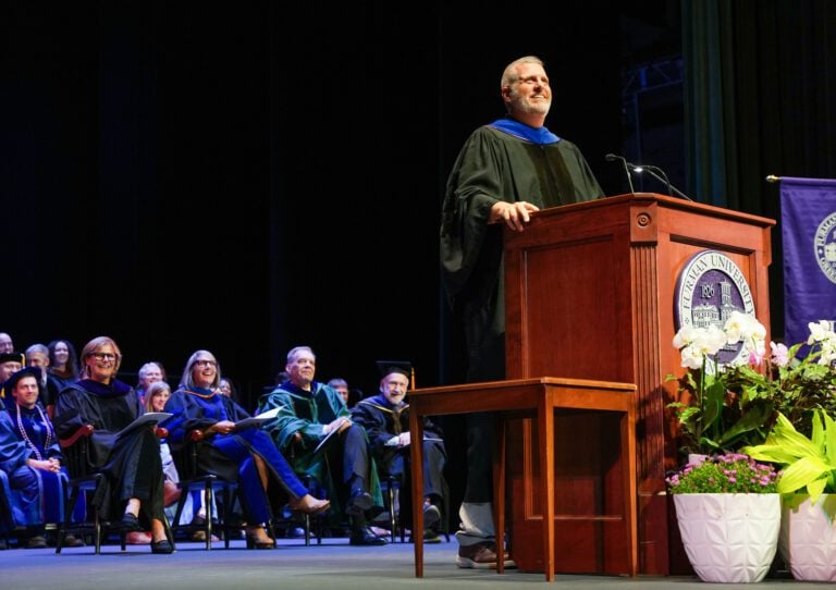 A man in an academic robe stands at a lectern and addresses a crowd out of frame during Furman University's Convocation.
