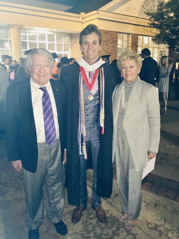 A younger male in a graduation gown stands between an older male and older female.