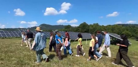 A group of people walk in a field containing solar panels, a solar farm.