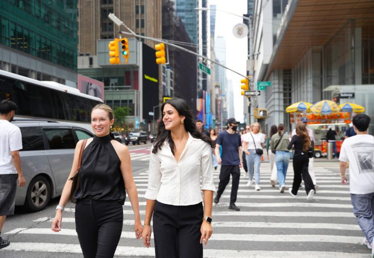 Two female college students cross the street at busy crosswalk with tall buildings surrounding them.