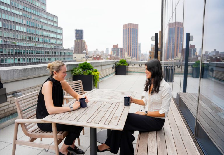 Two female college students sit on a patio at a table holding coffee mugs. A city skyline is in the background.