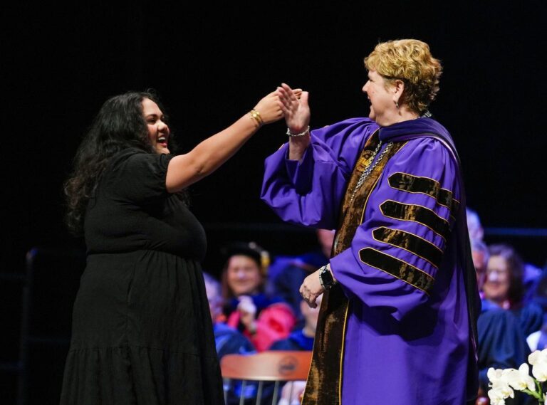 A young woman in a black dress "daps" an older woman in a purple ceremonial academic robe on stage at Furman University's indoors Convocation ceremony