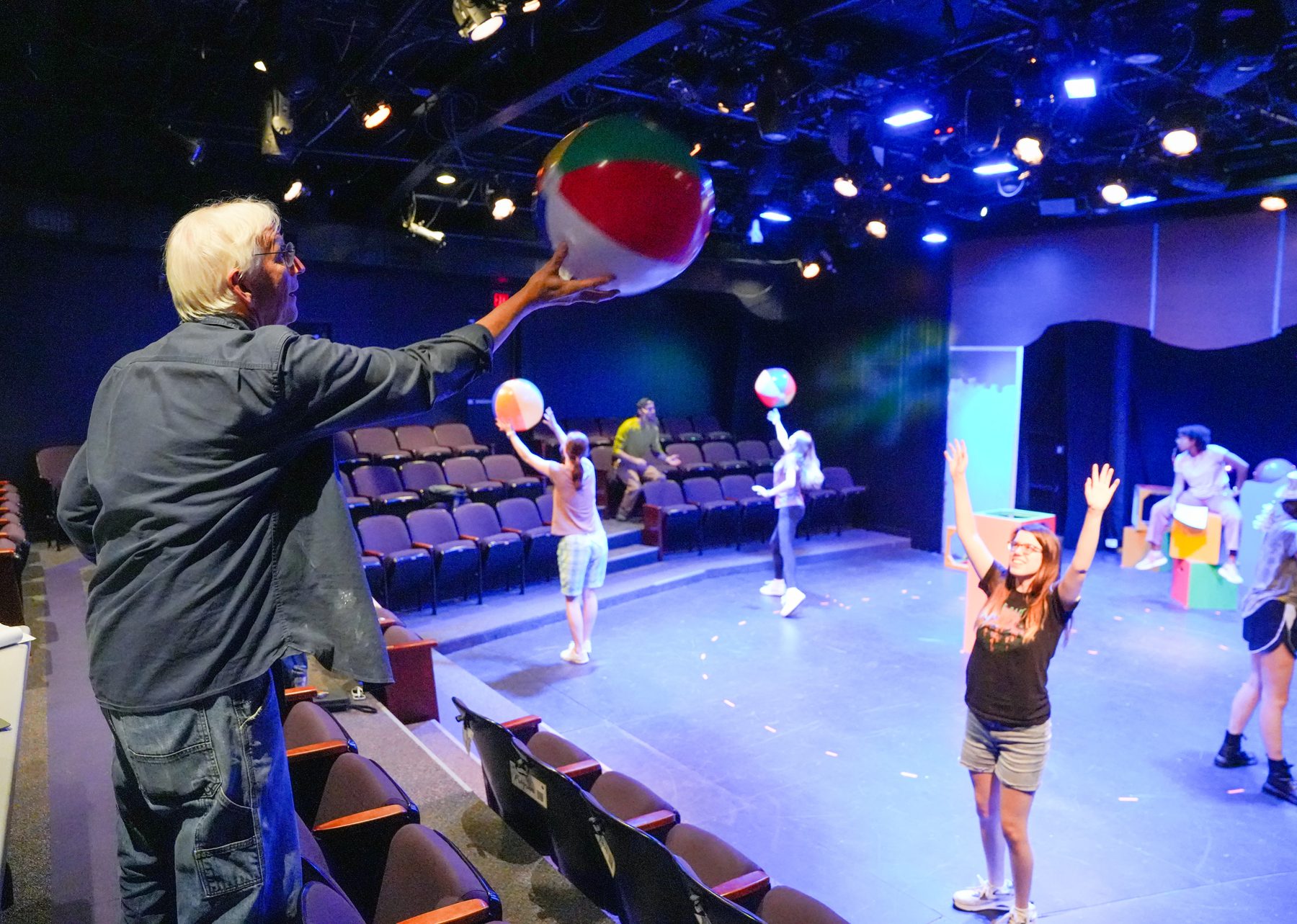 A man with white hair directs a theater of actors dressed as clowns.