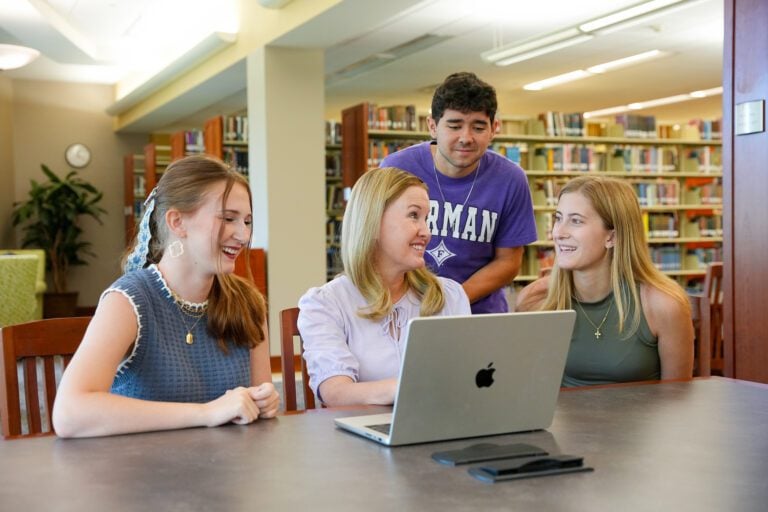 Three young students huddle around a Furman University staff member who sits behind a laptop explaining something to the students.