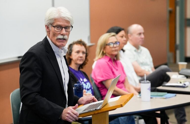 An older white man stands behind a lectern looking at a student out of frame while a panel of professors sit behind him during a discussion.