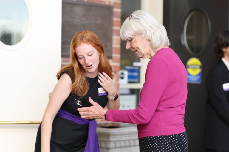 A young white woman leans down to look at something on a cellphone being held by an older woman.