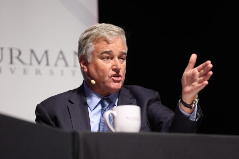 An older white man sits behind a table, gesturing with his left hand as he speaks to an unseen crowd.