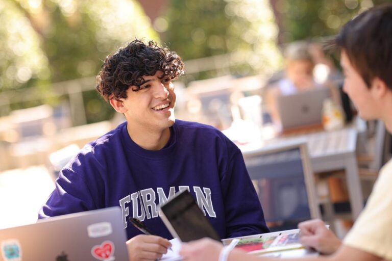 A young man sits in a purple sweater, smiling at an outdoor table speaking with another person who is slightly out of frame.