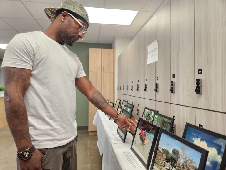 A Black man reaches down in an indoor space to reach out to one of several framed images at an art exhibition of photographs he and other people took.