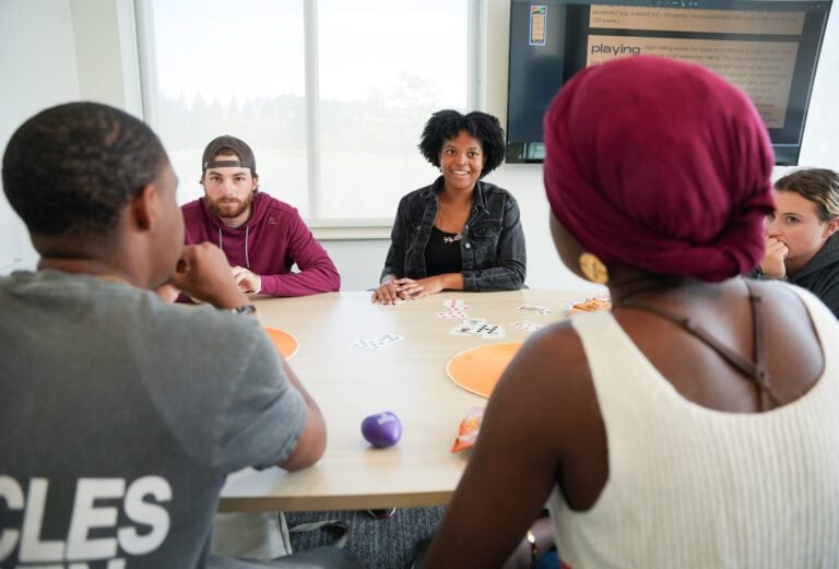Three female and two male students play a card game at a table.