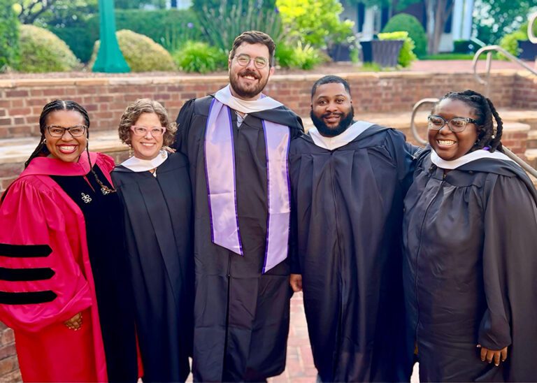 Three women and two men wearing graduation gowns stand together outside. 