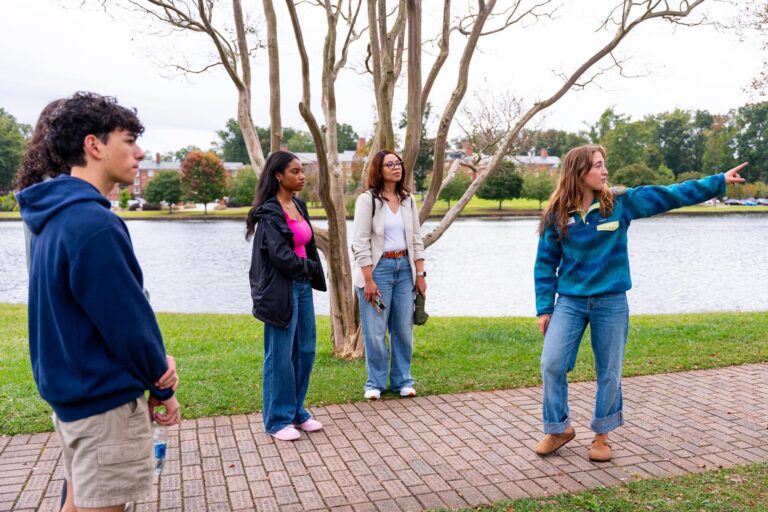 A young woman points off screen as she leads a tour group on a college campus