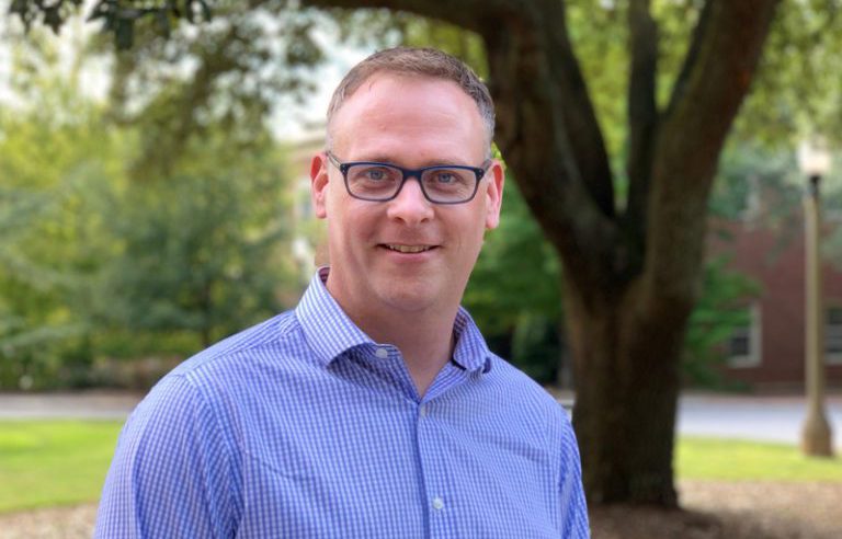 A man in glasses stands outside wtih a tree in the background.