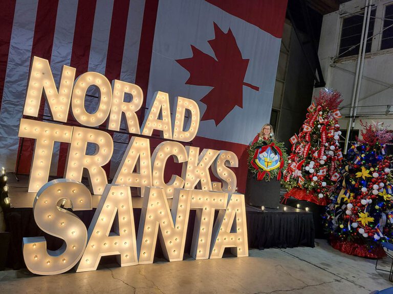 A woman stands behind a podium next to a large, lighted sign that reads NORAD Tracks Santa.