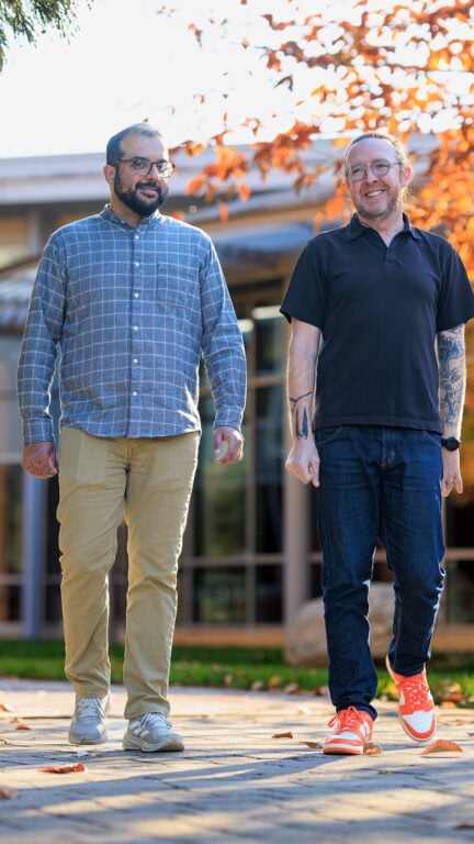 Two men dressed casually walk outside with fall leaves behind them. 