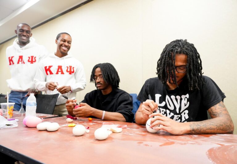 Four Black men make stress balls as a craft service project, wearing shirts bearing the logo of the Kappa Alpha Psi fraternity.