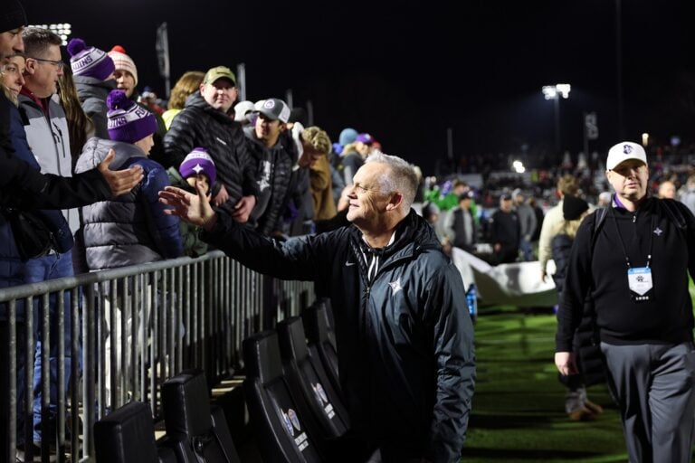 A white man wearing a black jacket greets fans at a soccer stadium. Doug Allison.
